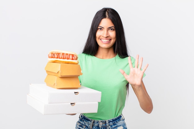 Mooie Spaanse vrouw die lacht en er vriendelijk uitziet, nummer vijf laat zien en fastfooddozen vasthoudt