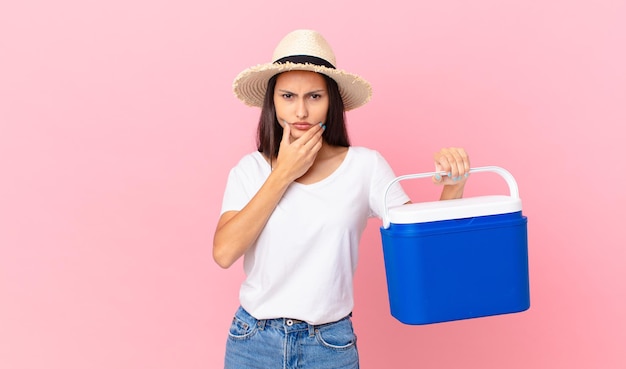 Mooie Spaanse vrouw met mond en ogen wijd open en hand op kin en met een draagbare koelkast