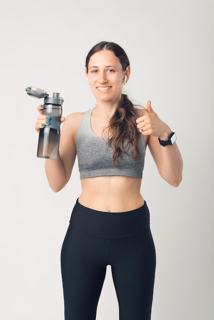 Mooie sportieve vrouw die drinkfles water gebruikt, glimlacht, duim omhoog doet uitstekend en goedkeuringsteken