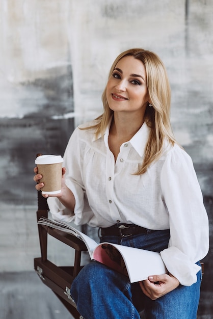 Mooie stijlvolle blonde vrouw in casual denim kleding en een witte blouse met volumineuze mouwen met een tijdschrift en koffie in haar handen. Zachte selectieve aandacht.