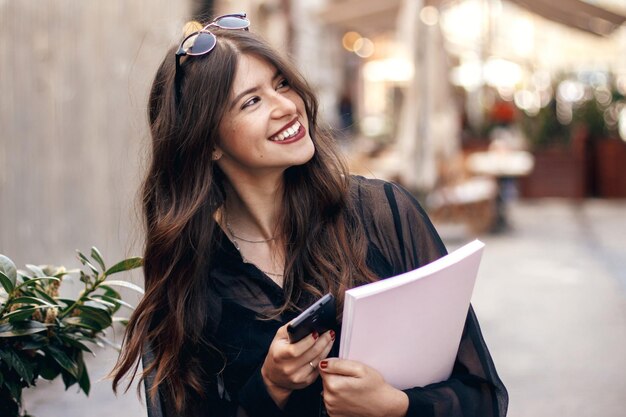 Mooie stijlvolle hipster meisje met telefoon en tijdschrift lopend en lachend in zonnige straat gelukkige prachtige vrouw gekleed in modieuze outfit genietend van tijd in Europese stad