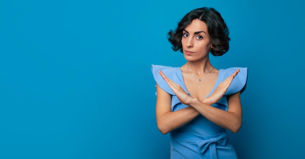 Mooie stijlvolle serieuze jonge vrouw in een blauwe jurk toont gekruiste handen in de camera geïsoleerd op blauwe achtergrond