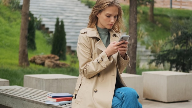 Mooie stijlvolle student meisje chatten met vriend op telefoon zittend op een bankje met boeken in park Studeer buiten concept