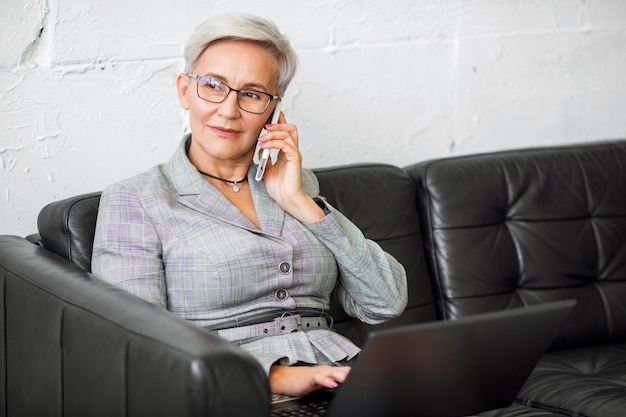 mooie stijlvolle volwassen vrouw in pak zittend op de bank met laptop en telefoon