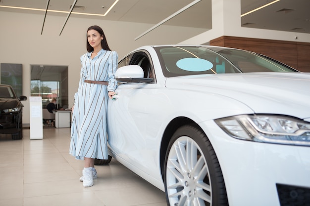 Mooie stijlvolle vrouw die in de buurt van haar nieuwe auto staat bij de autodealer, kopieer ruimte