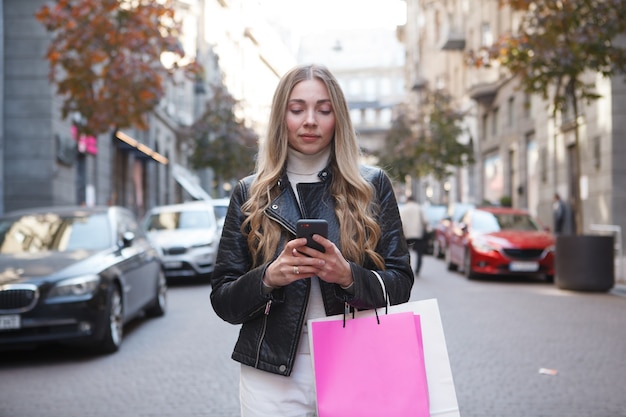 Mooie stijlvolle vrouw met boodschappentassen die sms't op haar smartphone die op straat staat