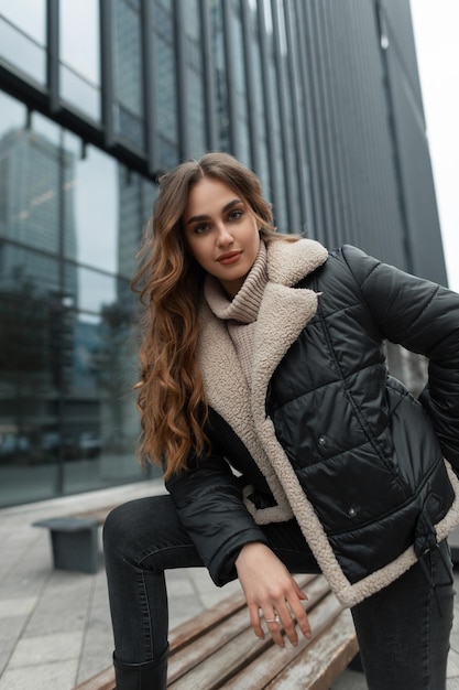 Mooie stijlvolle vrouw met een krullend kapsel in een modieuze winteroutfit met een modieuze jas van gebreide trui en spijkerbroek staat en poseert in de stad