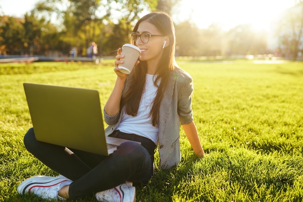 Mooie stijlvolle vrouw zittend op groen gras met laptop en telefoon in de handen Lifestyle concept