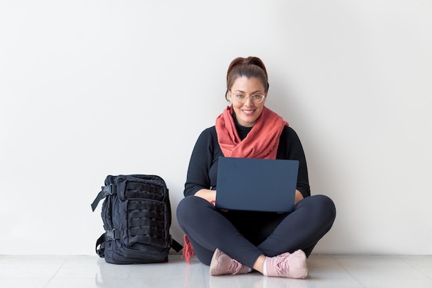 Foto mooie student die glimlacht tijdens het gebruik van laptop op de vloer met zwarte bagage rode sjaal en bril