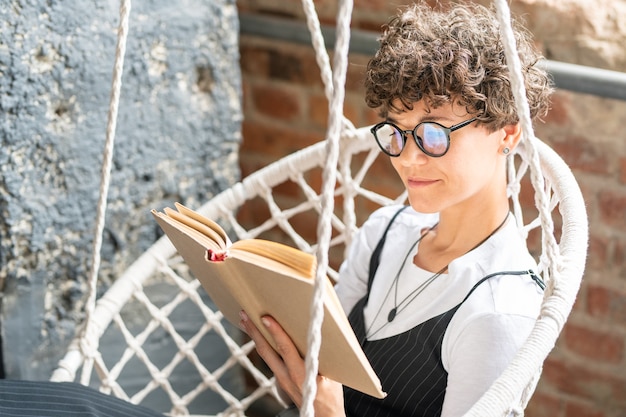 Mooie student in brillen lezen boek thuis zittend in hangende fauteuil op uw gemak