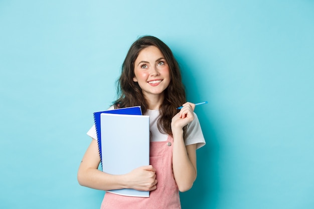 Mooie studente die nadenkend kijkt naar de bovenste kopieerruimte, staande met notebooks huiswerk en peinzend lachend, staande tegen een blauwe achtergrond.