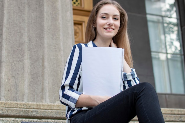 Mooie studente zit op de trap in de buurt van de universiteit met een rugzak met papieren in haar handen en kijkt omhoog, ze leert lessen tijdens de pauze