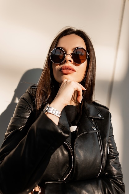 Mooie swag vrouw met natuurlijke grote lippen in mode leren jas met vintage ronde zonnebril poseert op straat in zonlicht