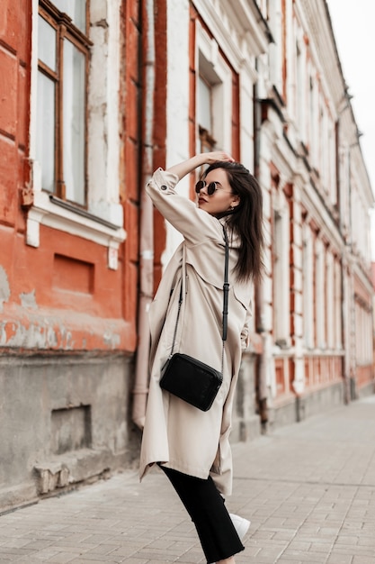 Mooie trendy jonge vrouw in vintage zonnebrillen in voorjaar stijlvolle trenchcoat met lederen zwarte handtas geniet van een wandeling langs de straat. aantrekkelijk meisje mannequin in modieuze kleding poses in de stad.