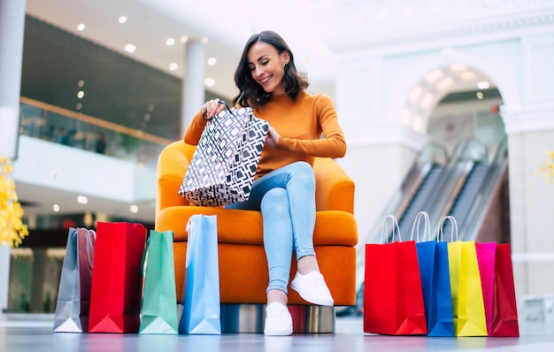 Mooie trendy jonge vrouw met veel kleurrijke boodschappentassen in goed humeur met slimme telefoon en creditcard zittend in het winkelcentrum tijdens zwarte vrijdag