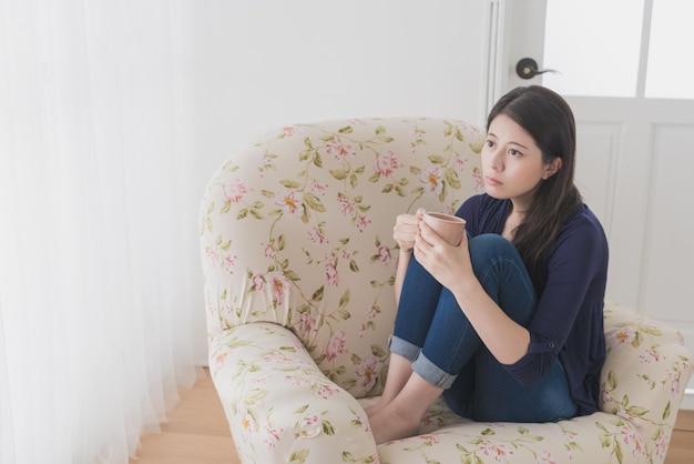 Mooie trieste vrouw die zich depressief voelt terwijl ze thuis naar het raam kijkt en een warme koffiekop vasthoudt terwijl ze op de bank in de woonkamer zit te dagdromen.