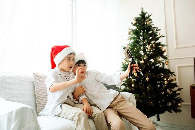 Mooie twee broers die thuis een selfie met mobiele telefoon nemen. Gelukkige jonge geitjes die bij de kerstboom zitten.