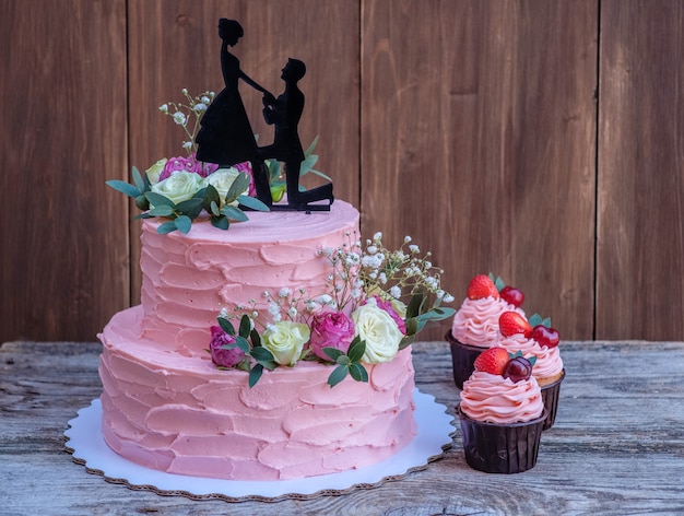 Mooie tweelaagse bruidstaart met roze kaascrème, versierd met levende rozen en een figuur van een verliefd stel, op een houten tafel met cupcakes