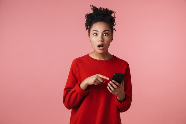 mooie verbazingwekkende geschokt opgewonden jonge afrikaanse vrouw poseren geïsoleerd over roze muur met behulp van mobiele telefoon.