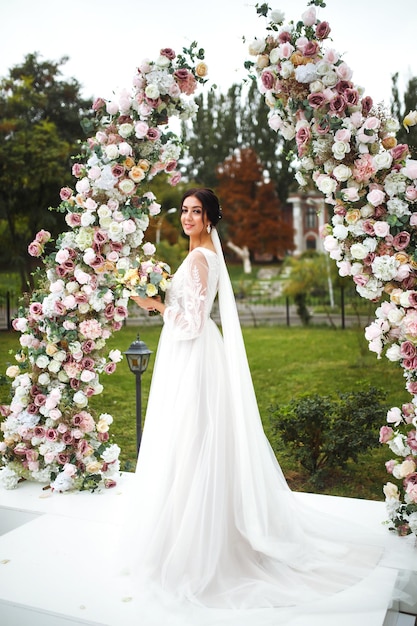 Mooie verloofde in witte trouwjurk met boeket staan in de buurt van de huwelijksboog van bloemen