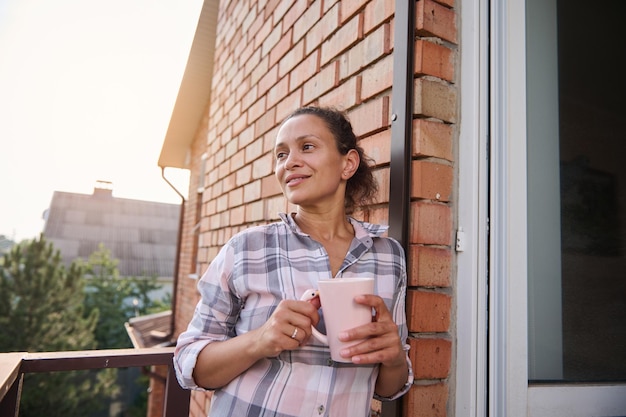 Mooie verrukkelijke vrouw die een kop warme drank vasthoudt en geniet van haar vrije dag op het balkon van het plattelandsresort