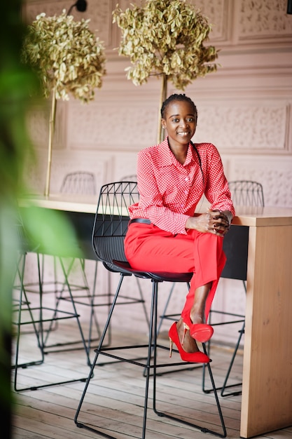 Mooie vlechten zakelijke afro-amerikaanse dame heldere bazige persoon vriendelijke slijtage kantoor rood shirt en broek, zittend op een stoel.