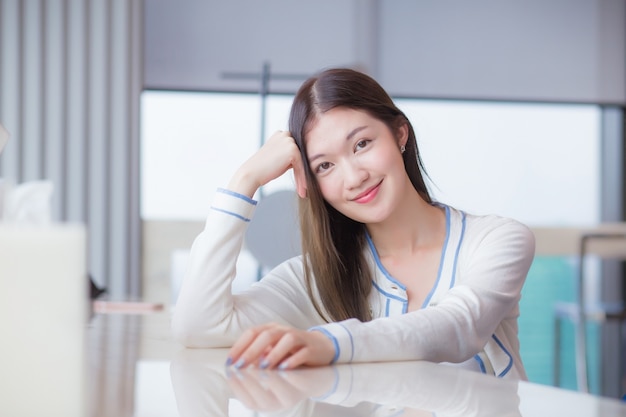 Mooie volwassen leeftijd Aziatische professionele vrouw met lang haar in wit overhemd kijkt naar de camera die haar gelukkig glimlacht terwijl ze op een stoel zit in het kantoor van de werkplek met een glazen gebouw als achtergrond.
