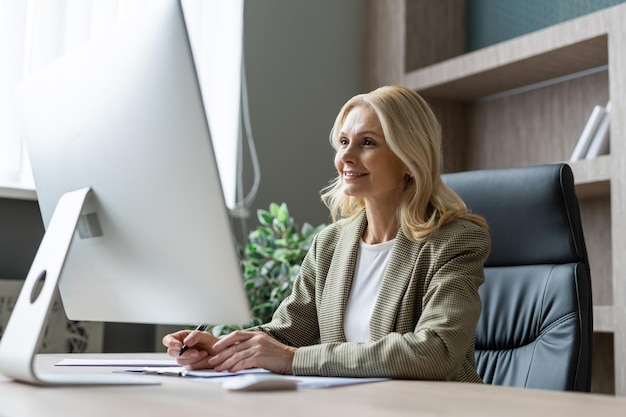 Mooie volwassen volwassen zakenvrouw met elegante jurk zittend aan een computerbureau op kantoor