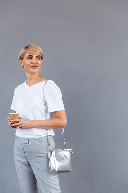 Mooie volwassen vrouw die witte t-shirt draagt die zich geïsoleerd over grijze muur bevindt, en afhaalkoffie houdt