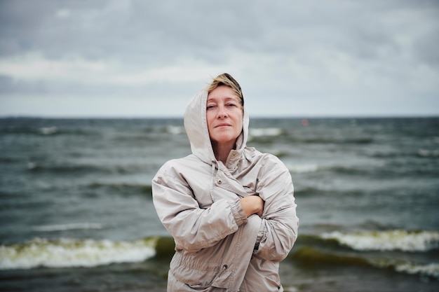 Mooie volwassen vrouw in een capuchon en gewikkeld in een jas tegen de achtergrond van een stormachtige zee