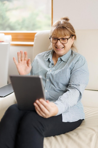 Mooie volwassen vrouw met bril gebruikt een digitale tablet en glimlacht terwijl ze thuis op de bank zit