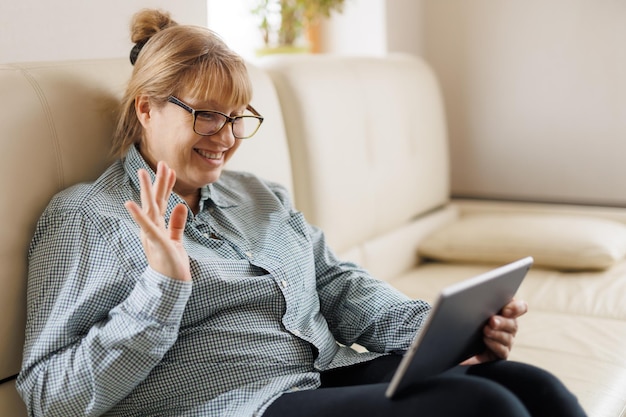 Mooie volwassen vrouw met bril gebruikt een digitale tablet en glimlacht terwijl ze thuis op de bank zit