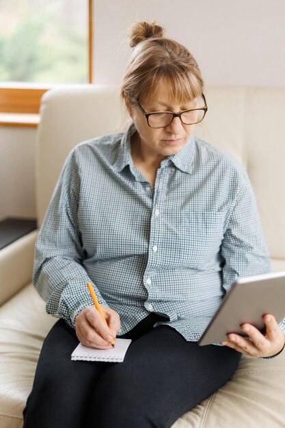 Mooie volwassen vrouw met bril gebruikt een digitale tablet en glimlacht terwijl ze thuis op de bank zit