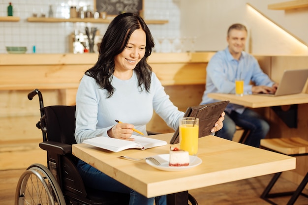 Mooie vrolijke gehandicapte vrouw zittend in een rolstoel en schrijven in haar notitieblok en werken op haar tablet in een café en een man zit op de achtergrond