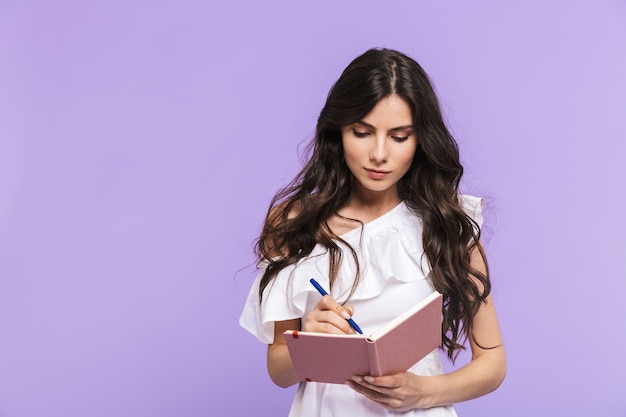 Mooie vrolijke jonge vrouw die een zomeroutfit draagt, geïsoleerd over een violette muur en aantekeningen maakt in haar dagboek