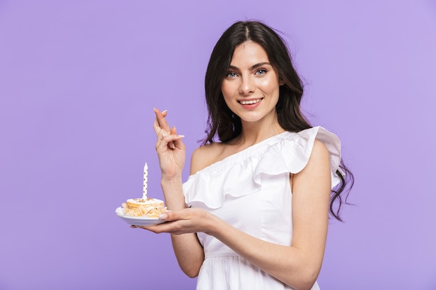 Mooie vrolijke jonge vrouw die een zomeroutfit draagt, geïsoleerd over een violette muur, verjaardag viert met cake