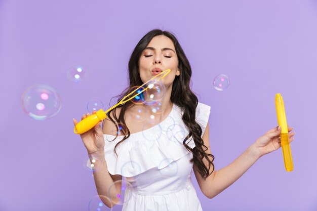 Mooie vrolijke jonge vrouw die een zomeroutfit draagt, geïsoleerd over een violette muur, zeepbellen blazend