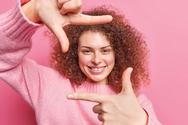 Foto mooie vrolijke jonge vrouw die handgebaar maakt glimlacht gelukkig draagt casual trui maakt geschoten met vingers foto's iets in gedachten creats meesterwerk staat over roze muur.