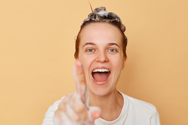 Foto mooie vrolijke vrolijke vrouw wast haar past shampoo toe en neemt een douche geïsoleerd over beige achtergrond met schuim op haar vinger klik op onzichtbare knop