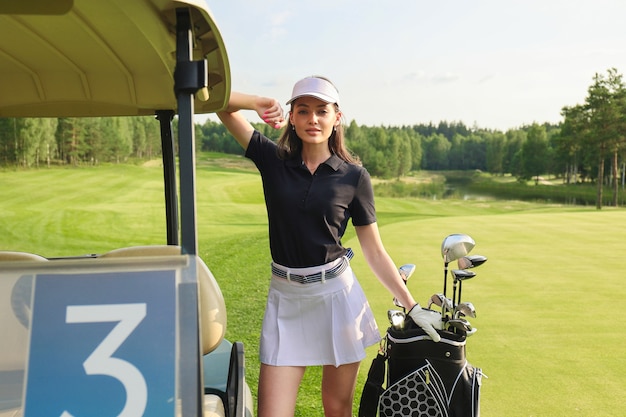 Mooie vrolijke vrouw die zich dichtbij in golfkar bevindt met golr-uitrusting.