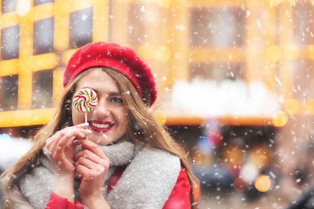 Mooie vrolijke vrouw gekleed in een rode jas en een warme sjaal met karamelsnoep tijdens de sneeuwval