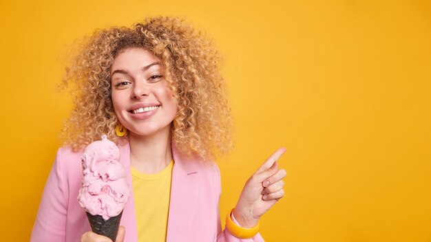 Mooie vrolijke vrouw met krullend haar lacht tandjes geeft aan dat in de rechterbovenhoek ruimte is voor uw advertentie, promoot smakelijke zomerdessert poses met ijs tegen gele muur