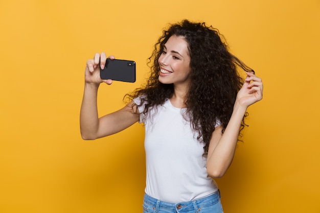 mooie vrouw 20s met krullend haar lachen en selfie foto nemen op mobiele telefoon geïsoleerd op geel