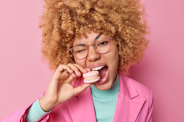 Mooie vrouw bijt smakelijke macaron geniet van zoet eten heeft suikerverslaving houdt mond wijd open draagt bril en elegante modieuze jas geïsoleerd over roze studiomuur Brekend dieet