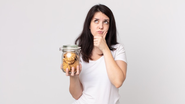 Mooie vrouw denkt, voelt zich twijfelachtig en verward en houdt een glazen fles met koekjes vast