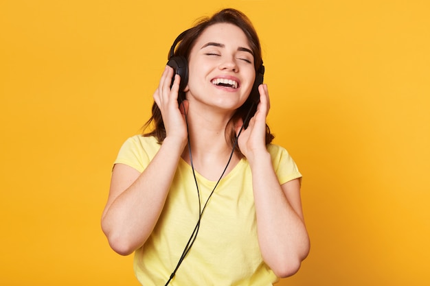 Mooie vrouw die aan muziek op gele muur luistert. Charmante dame poseren met gesloten ogen, geniet van het luisteren naar favoriete muziek, houdt een koptelefoon vast, zingt en ontspant. Levensstijl concept.