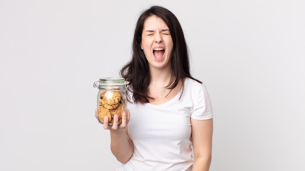 Mooie vrouw die agressief schreeuwt, erg boos kijkt en een glazen fles met koekjes vasthoudt