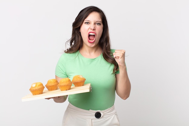 Mooie vrouw die agressief schreeuwt met een boze uitdrukking en een dienblad met muffins vasthoudt