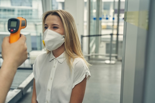 Mooie vrouw die een beschermend masker draagt terwijl ze tegenover de werknemer van de luchthaven staat en de temperatuur controleert