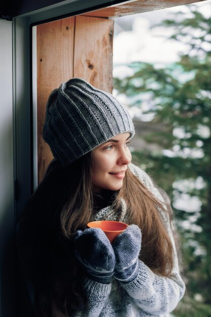 Mooie vrouw die een kopje koffie of cacao vasthoudt en drinkt in handschoenen die thuis bij het raam zitten